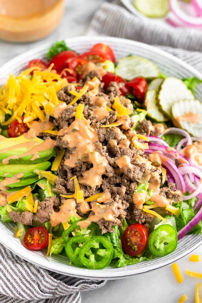 Burger salad filled with lettuce, ground beef, tomatoes, pickles, onions, avocado, cheese, and topped with special sauce. Behind it is a jar of special sauce and a plate of more toppings.