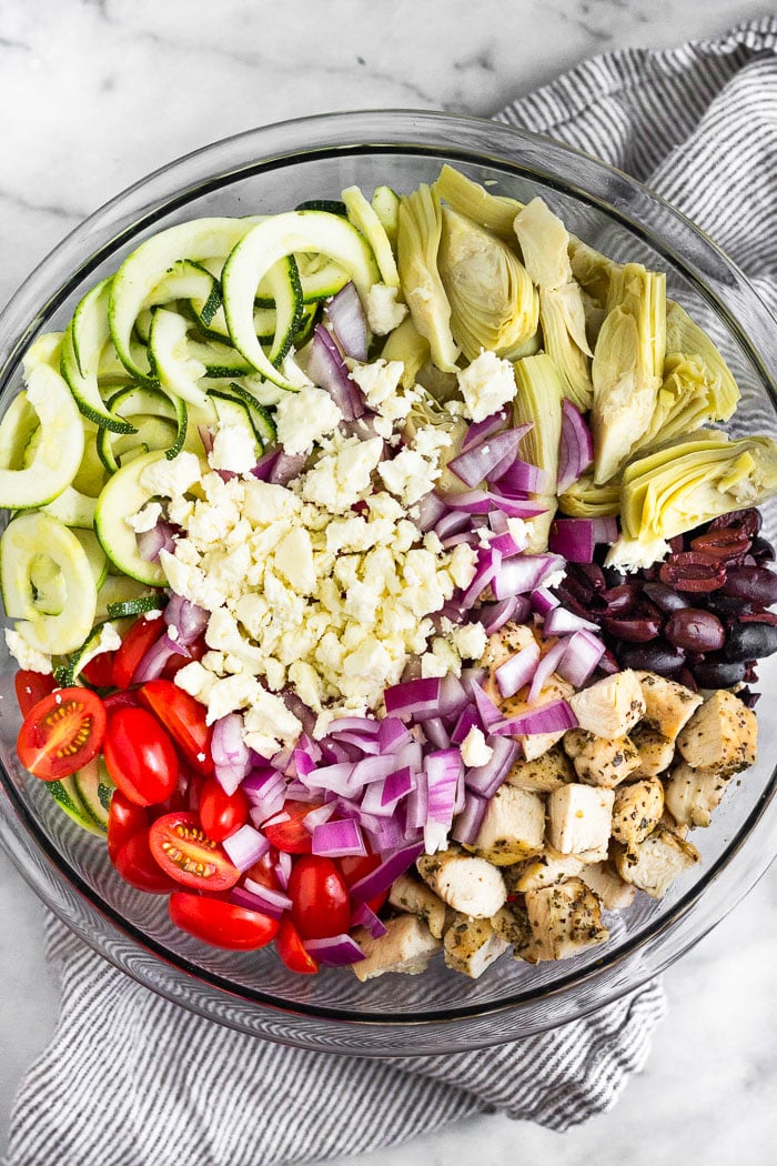 Bowl filled with chopped chicken, cherry tomatoes, zucchini noodles, artichoke hearts, olives, red onion, and feta cheese.