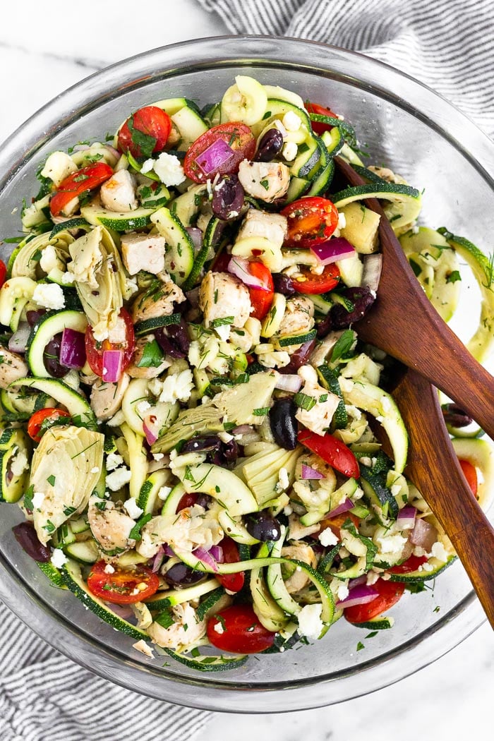 Bowl filled with greek zucchini noodle pasta salad with two wooden tongs in it.