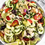 Greek chicken pasta salad with zucchini noodles in a large bowl. It is sitting on a stripped towel surrounded by lemon wedges, fresh herbs, and a spoon.