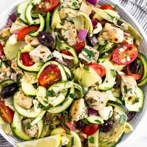 Greek chicken pasta salad with zucchini noodles in a large bowl. It is sitting on a stripped towel surrounded by lemon wedges, fresh herbs, and a spoon.