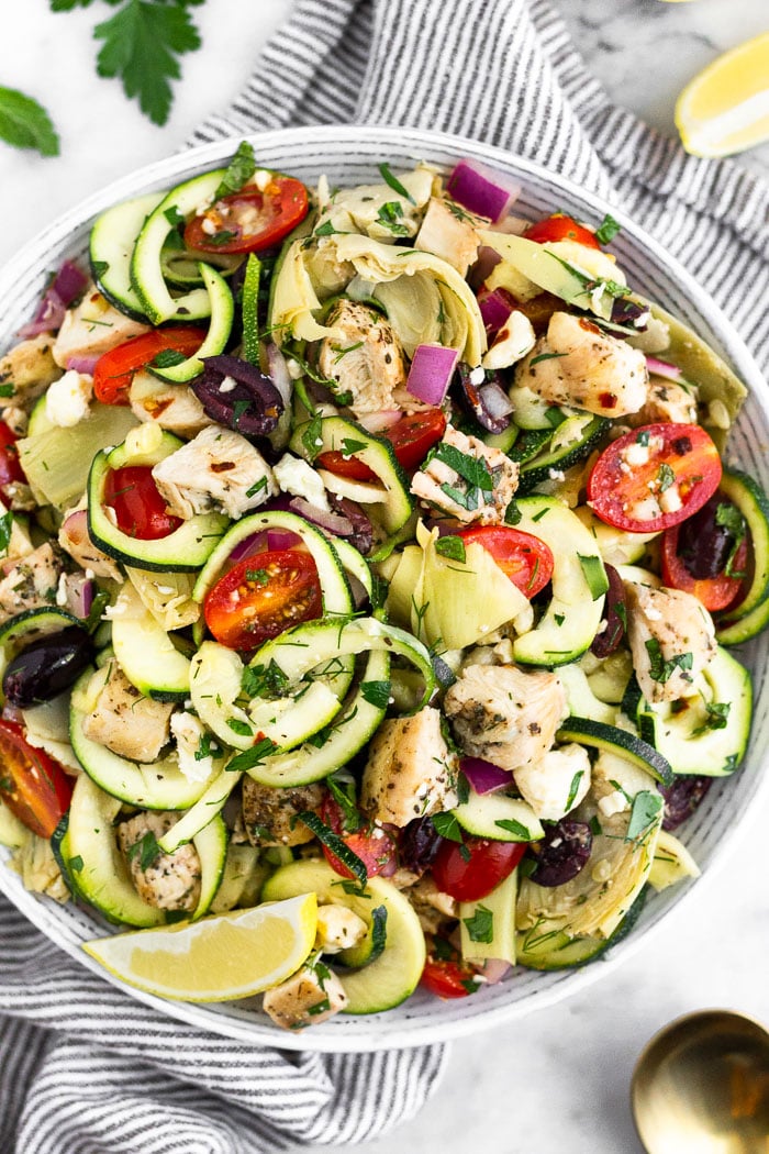 Greek chicken pasta salad with zucchini noodles in a large bowl. It is sitting on a stripped towel surrounded by lemon wedges, fresh herbs, and a spoon.