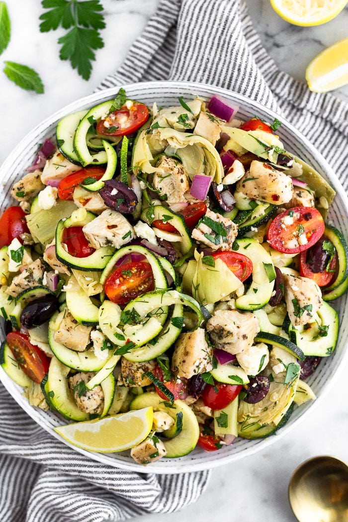Overhead shot of greek chicken pastas salad in a bowl with a lemon wedge on the side. The bowl is sitting on a towel surrounded by fresh herbs, lemon wedges, and a spoon.