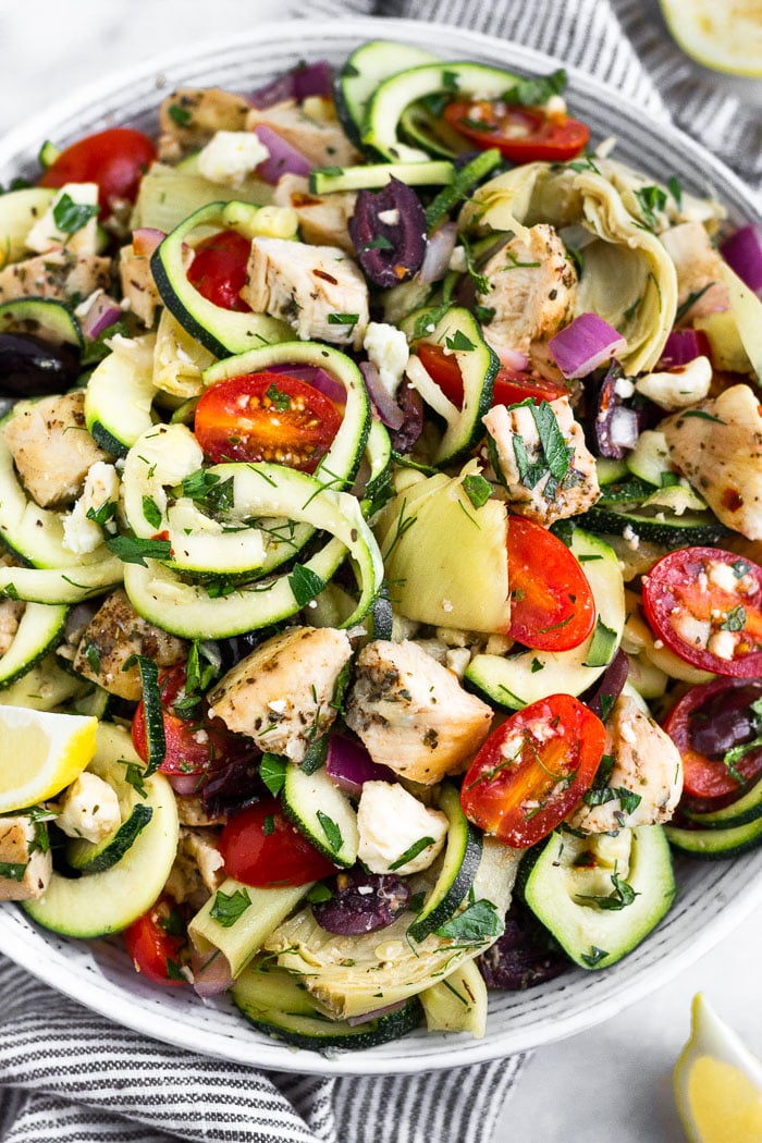 Close up a greek pasta salad with chicken and zucchini noodles in a large bowl. Lemon wedges are around the bowl.