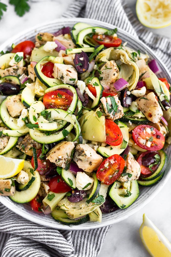 Greek chicken pasta salad filled with zucchini noodles. chicken, tomatoes, olives, red onion. artichokes, and fresh herbs. It is garnished with lemon wedges with a few more wedges around the bowl.