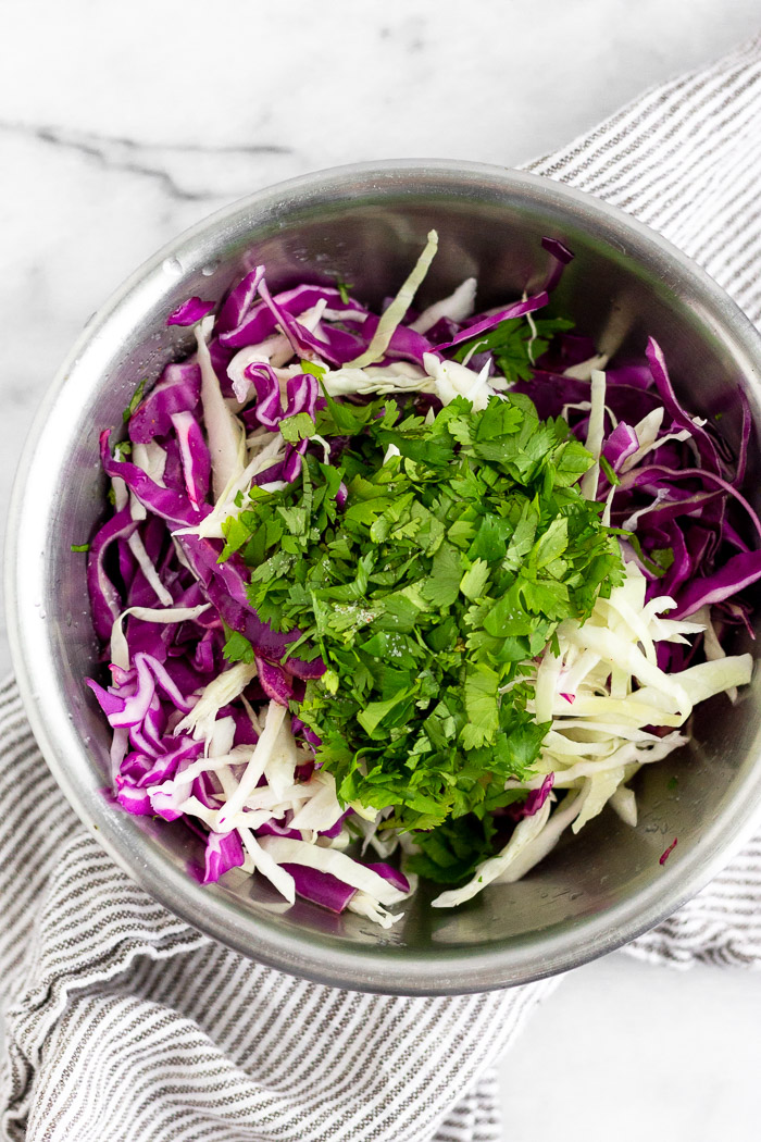 Bowl of shredded purple and green cabbage and chopped cilantro. Under the bowl is stripped towel.