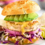 Mahi Mahi burger with shredded cabbage, pickled onions, chipotle mayo, and avocado in a bun on a sheet plate. It is surrounded by chips, a lime wedge, and a small bowl of salsa. In the distance is another burger and a bowl of shredded cabbage.