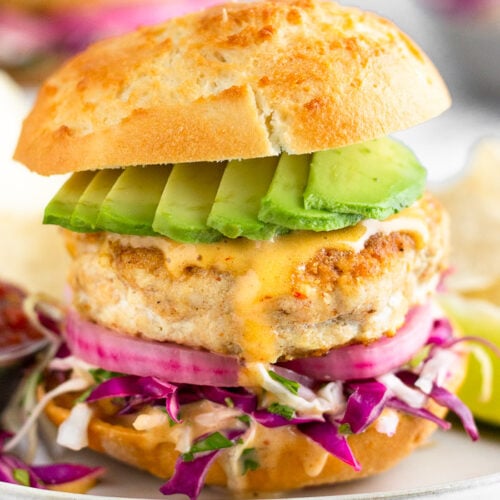 Mahi Mahi burger with shredded cabbage, pickled onions, chipotle mayo, and avocado in a bun on a sheet plate. It is surrounded by chips, a lime wedge, and a small bowl of salsa. In the distance is another burger and a bowl of shredded cabbage.