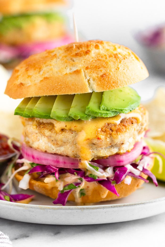 Mahi Mahi burger with shredded cabbage, pickled onions, chipotle mayo, and avocado in a bun on a sheet plate. It is surrounded by chips, a lime wedge, and a small bowl of salsa. In the distance is another burger and a bowl of shredded cabbage.