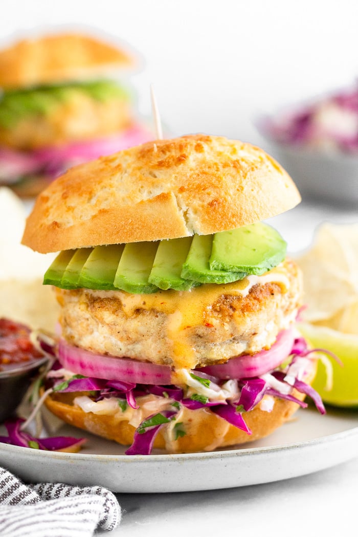 Fish burger in a bun stuffed with cabbage, pickled onions, chipotle mayo, and avocado with chips and a lime wedge next to it. Behind it is another burger and a bowl of cabbage.