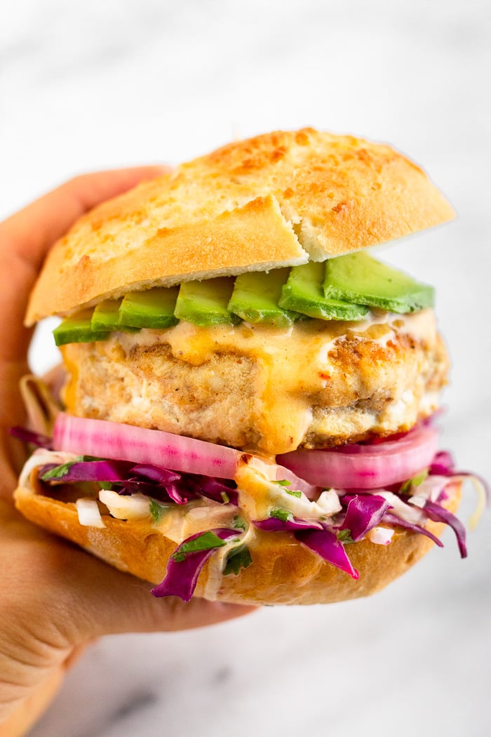 White background with a hand holding a Mahi Mahi burger with shredded cabbage, pickled onions, chipotle mayo, and sliced avocado in a bun