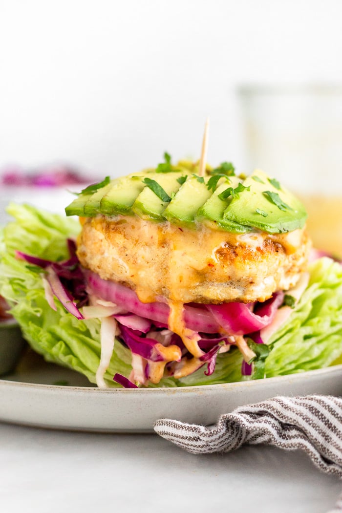Mahi Mahi burger on a lettuce bun with shredded cabbage, pickled onions, chipotle mayo, and chopped cilantro. In the distance behind it is a small bowl of shredded cabbage and a large jar of mayo.