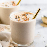Two tahini milkshakes in small glass jars. There are gold straws in them and they are topped with cinnamon and sesame seeds. There is towel and spoon around them.