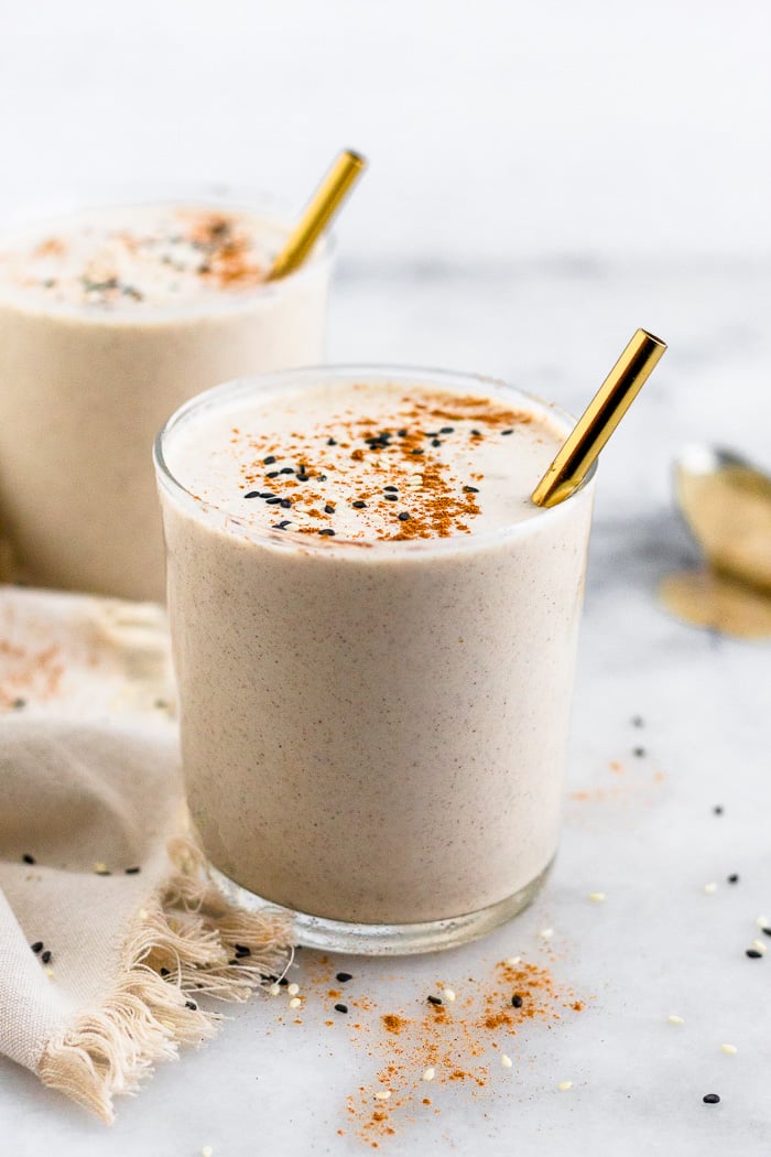 Two tahini milkshakes in small glass jars. There are gold straws in them and they are topped with cinnamon and sesame seeds. There is towel and spoon around them.