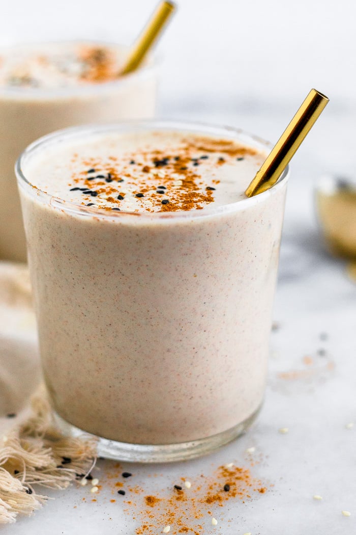 Cinnamon tahini milkshake with topped more cinnamon and a straw coming out of it. There is another milkshake behind it and cinnamon surrounding them.