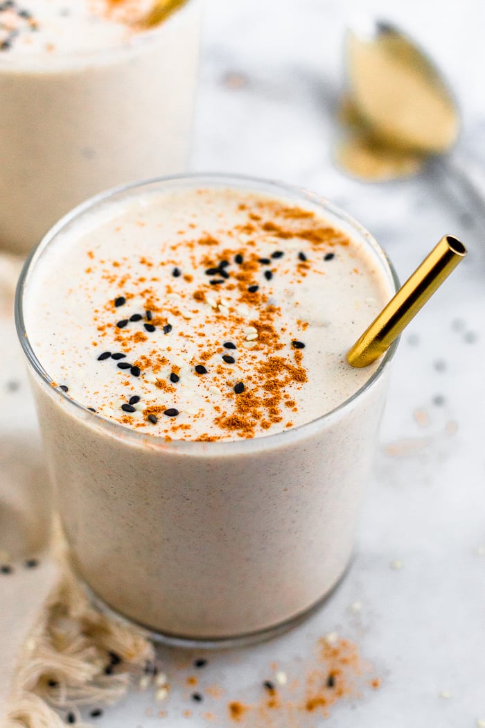 Close up of a cinnamon vanilla tahini milkshake topped with cinnamon and black and white sesame seeds. Surrounding it is more cinnamon, sesame seeds, and a tan linen.