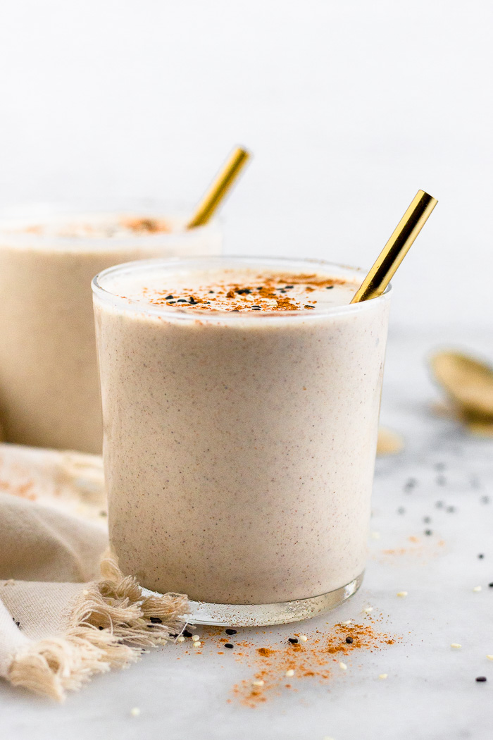 Two tahini milkshakes with gold straws coming out of them and topped with cinnamon and sesame seeds. One milkshake is in the distance with a spoon with tahini next to it.