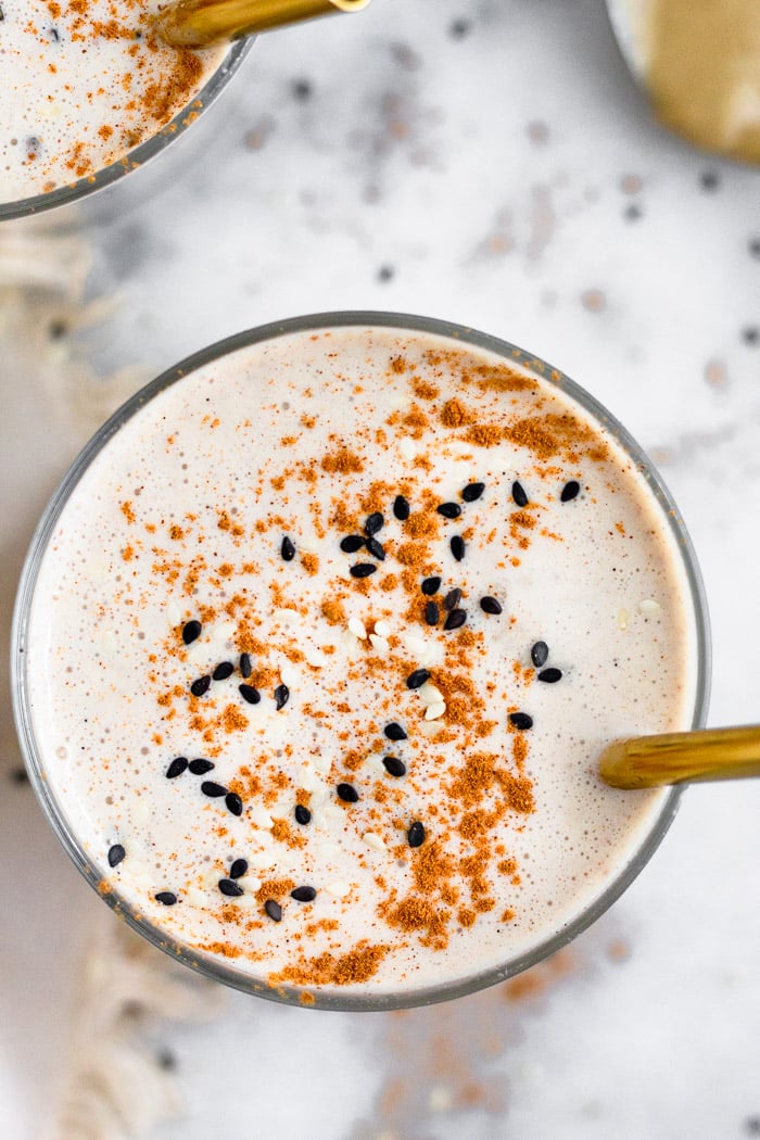 Overhead view of a milkshake topped with cinnamon and sesame seeds and a straw coming out of it. In the conversation is another milkshake and spoon with tahini.