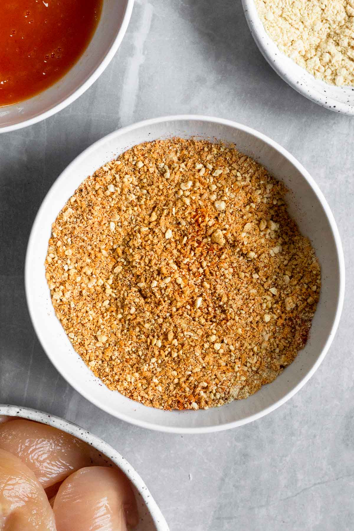 Bowl of ground up cashews, shredded, and spices. Next to it a bowl of whisked eggs, bowl of coconut flour, and a bowl of raw chicken tenderloins.