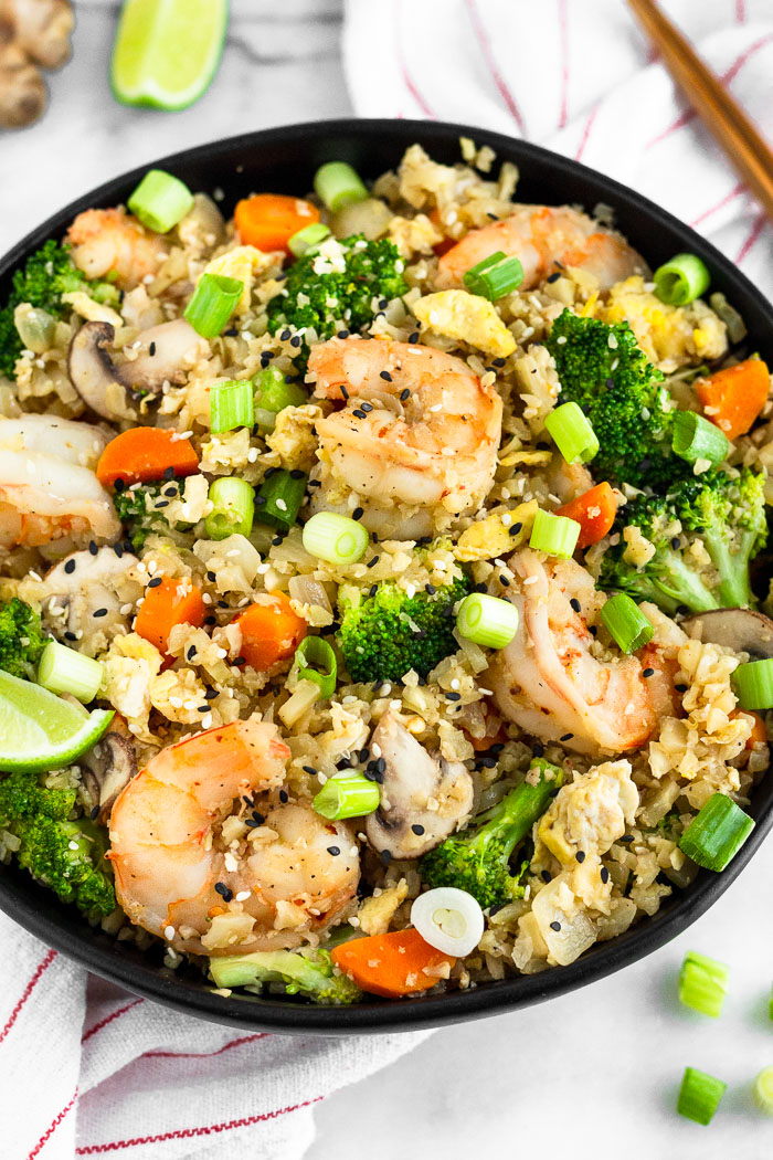 Bowl filled with Whole30 cauliflower shrimp fried rice sitting on top of a stripped towel with chopsticks, green onion, a lime wedge, and ginger around it.