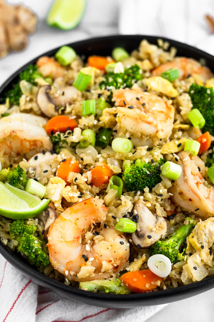Large black bowl filled with shrimp cauliflower fried rice garnished with a lime wedge. Behind it is another lime wedge and a piece of ginger.