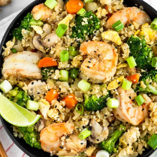 Large bowl of paleo cauliflower shrimp fried rice garnished with green onions and a lime wedge. The bowl is sitting on a stripped towel with chopsticks, ginger, a lime wedge, and green onions around it.