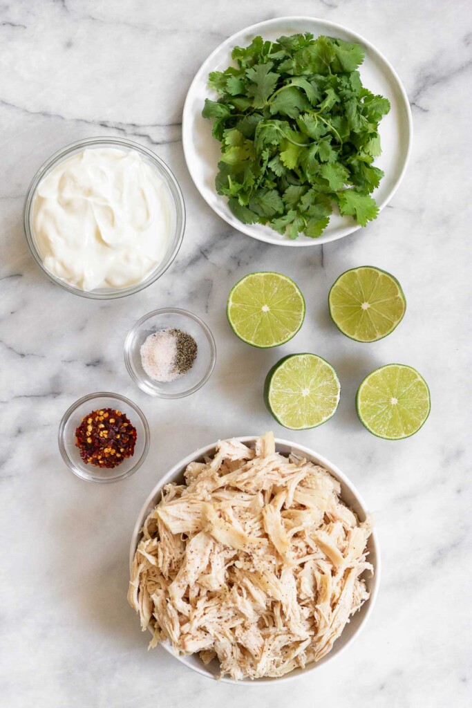 White marble counter with a plate of cilantro, 4 lime halves, a bowl of shredded chicken, a bowl of red pepper flakes, a bowl of salt and pepper, and a bowl of greek yogurt.