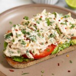 A piece of toast with mash avocado, sliced tomato, and cilantro lime chicken salad on top of it on a brown plate. Behind the plate is a small dish of cilantro and half a lime.