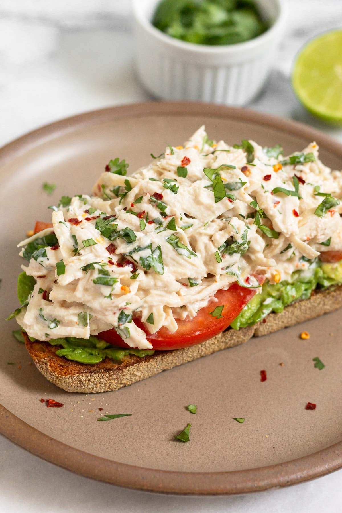 A piece of toast with mash avocado, sliced tomato, and cilantro lime chicken salad on top of it on a brown plate. Behind the plate is a small dish of cilantro and half a lime.