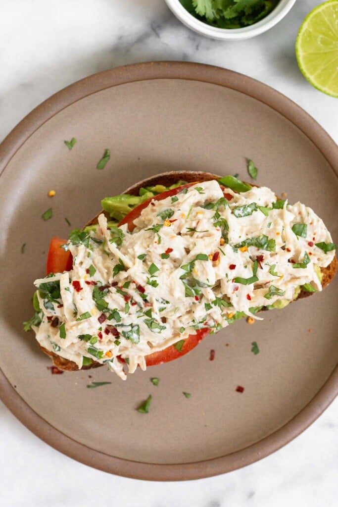 Overhead shot of chicken salad with greek yogurt, cilantro, and lime on top of sliced tomato and smashed avocado on a piece of toast on a plate.