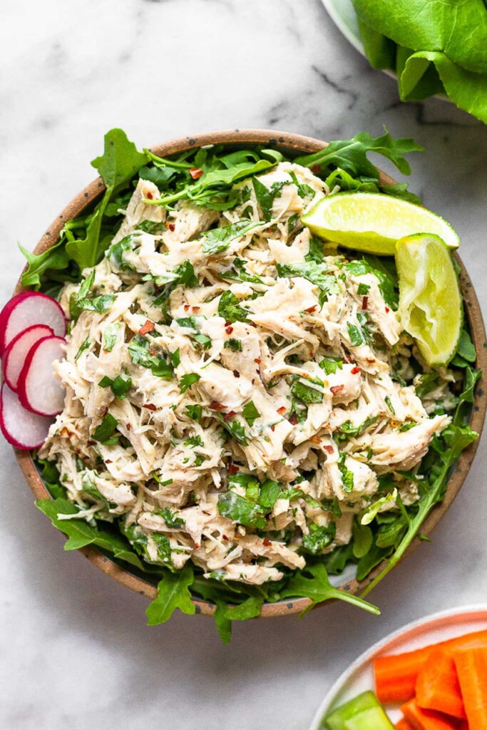 Cilantro lime chicken salad on top of some arugula in a bowl. It is garnished with lime wedges and sliced radishes. Next to the bowl is a plate carrots and a plate of lettuce leaves.