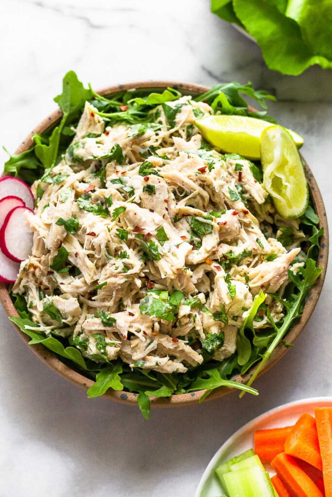 Chicken salad with cilantro and lime in a bowl overtop arugula. It is garnished with 2 lime wedges and sliced radishes. Next to the bowl is a plate of sliced carrots and celery and another plate of lettuce leaves.