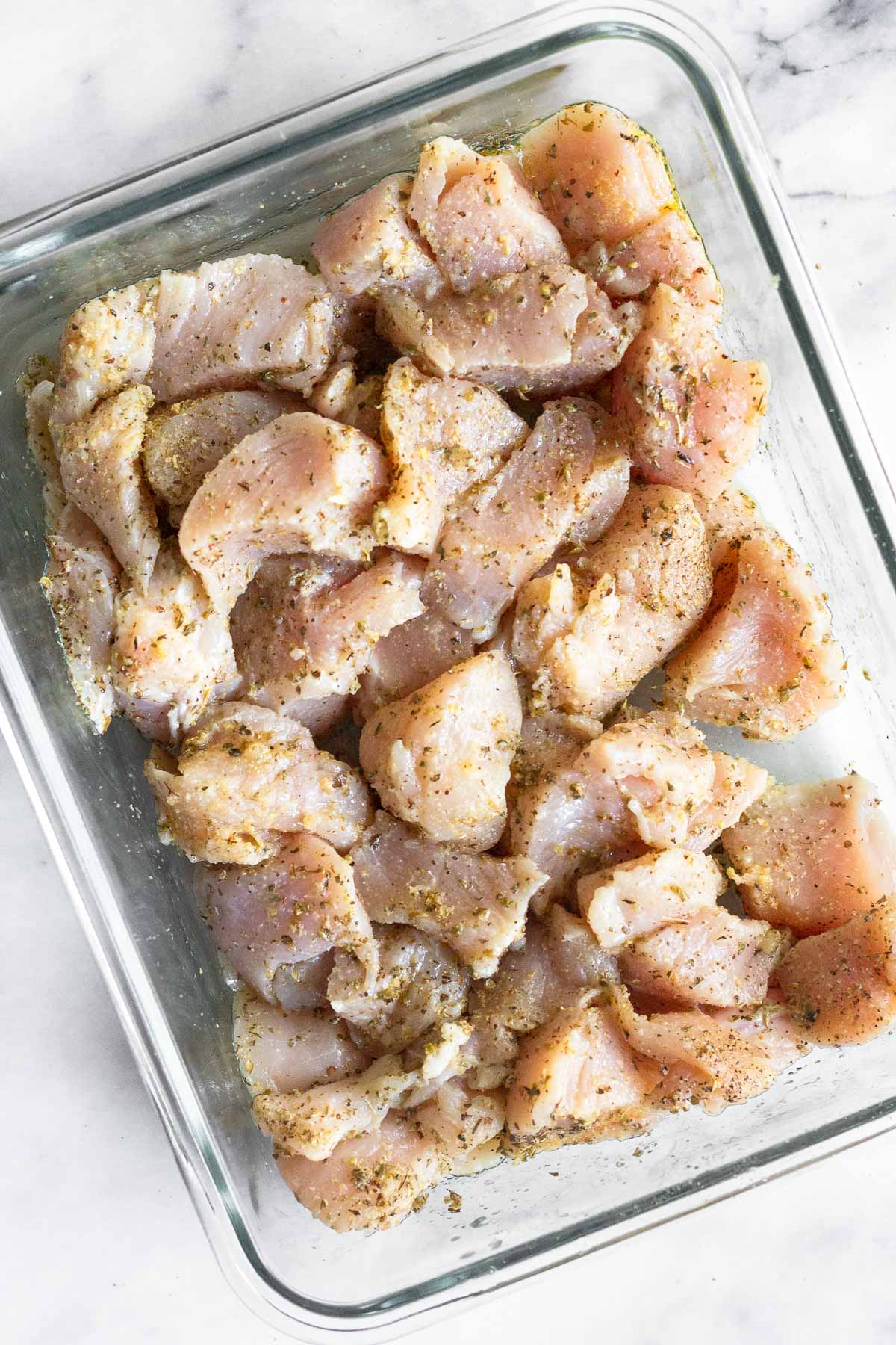 Glass container filled with cubed chicken marinating in oil and spices.