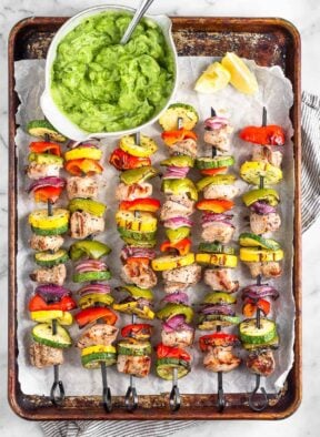 Grilled greek chicken kabobs on a baking sheet with a bowl of avocado tzatziki next to them.