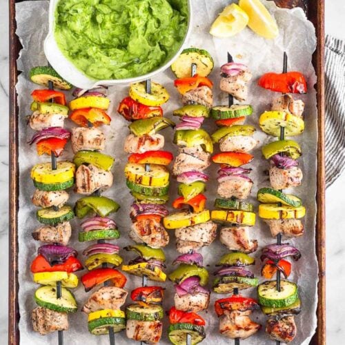 Grilled greek chicken kabobs on a baking sheet with a bowl of avocado tzatziki next to them.