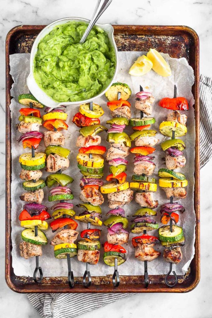 Grilled greek chicken kabobs on a baking sheet with a bowl of avocado tzatziki next to them.