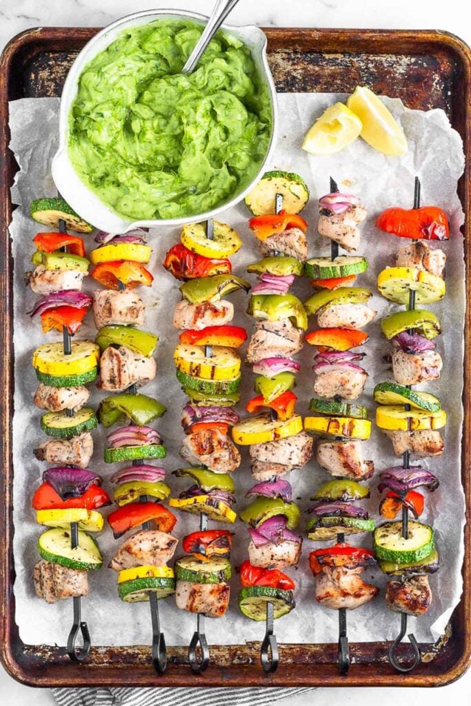 Greek chicken and veggie skewers on a baking sheet with lemon wedges and a bowl of avocado tzatziki.