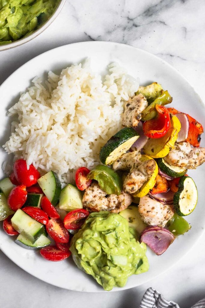 White plate with white rice, grilled chicken and veggies, avocado sauce, and a tomato and cucumber salad.