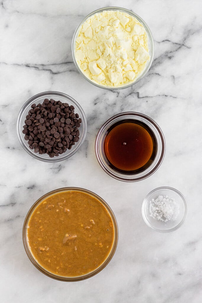 White marble counter with a bowl of protein powder, a bowl of maple syrup, a bowl of salt, a bowl of peanut butter, and a bowl of mini chocolate chips.