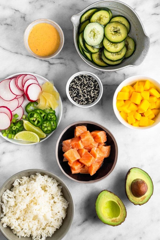 White counter with a bowl of sliced cucumbers, bowl of cubed mangos, bowl of cubed salmon, bowl of white rice, bowl of avocado, bowl of spicy mayo, and a bowl of sesame seeds. A plate is filled with radishes, jalapeños, pickled ginger, and lime wedges.