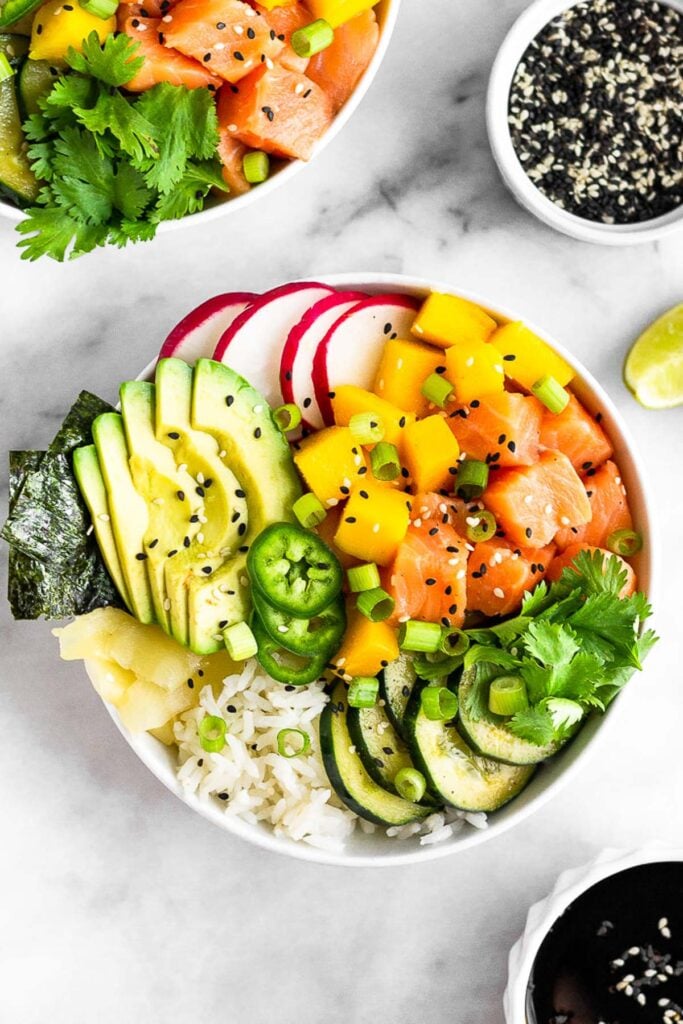 Overhead shoot of a Hawaiian poke bowl filled with salmon, rice, cucumbers, mango, avocado, radish, jalapenos, cilantro, pickled ginger, nori, green onion, and sesame seeds. It is surrounded by a bowl of soy sauce, lime wedges, sesame seeds, and another poke bowl.