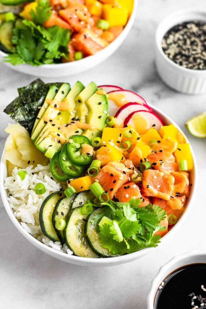 Spicy salmon poke bowl filled with white rice, cucumbers, salmon, mango, radishes, avocados, jalapeños, nori, and pickled ginger. It is garnished with cilantro, sesame seeds, and green onions and drizzled with spicy mayo. Behind it is another bowl, a bowl of sesame seeds, and lime wedges.
