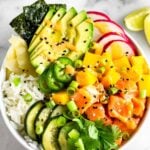 Salmon poke bowl with salmon, rice, veggies, mango, and avocado drizzled with spicy mayo. Behind it is another bowl, more sesame seeds, lime wedges, and a bowl of soy sauce is in front of it.