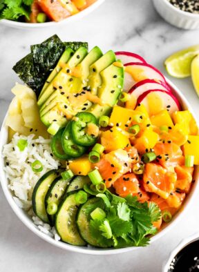 Salmon poke bowl with salmon, rice, veggies, mango, and avocado drizzled with spicy mayo. Behind it is another bowl, more sesame seeds, lime wedges, and a bowl of soy sauce is in front of it.