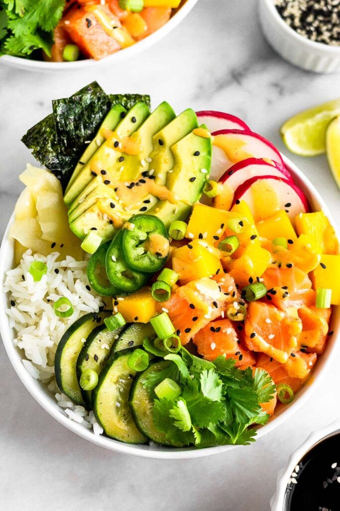 Salmon poke bowl with salmon, rice, veggies, mango, and avocado drizzled with spicy mayo. Behind it is another bowl, more sesame seeds, lime wedges, and a bowl of soy sauce is in front of it.