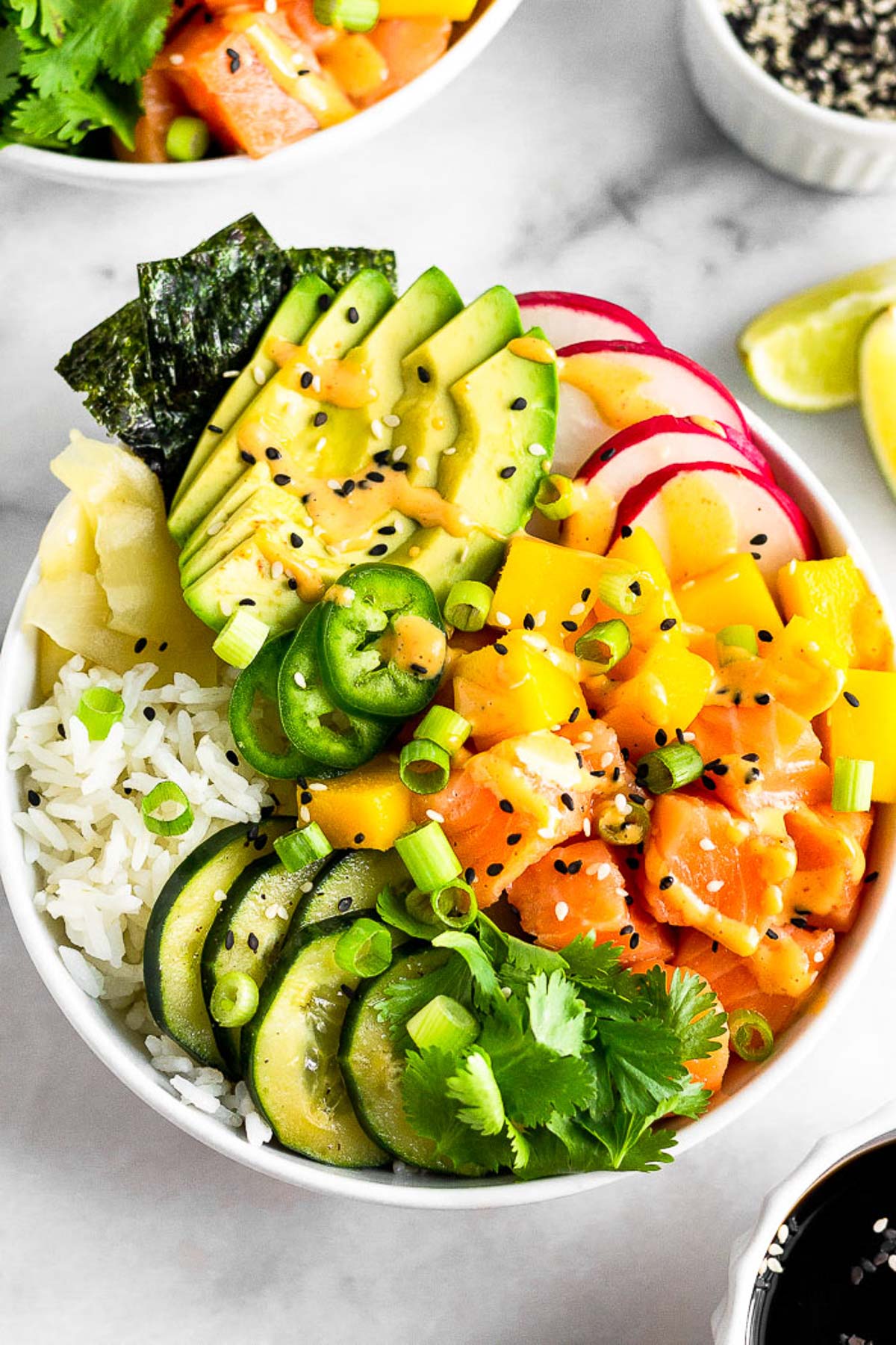 Salmon poke bowl with salmon, rice, veggies, mango, and avocado drizzled with spicy mayo. Behind it is another bowl, more sesame seeds, lime wedges, and a bowl of soy sauce is in front of it.