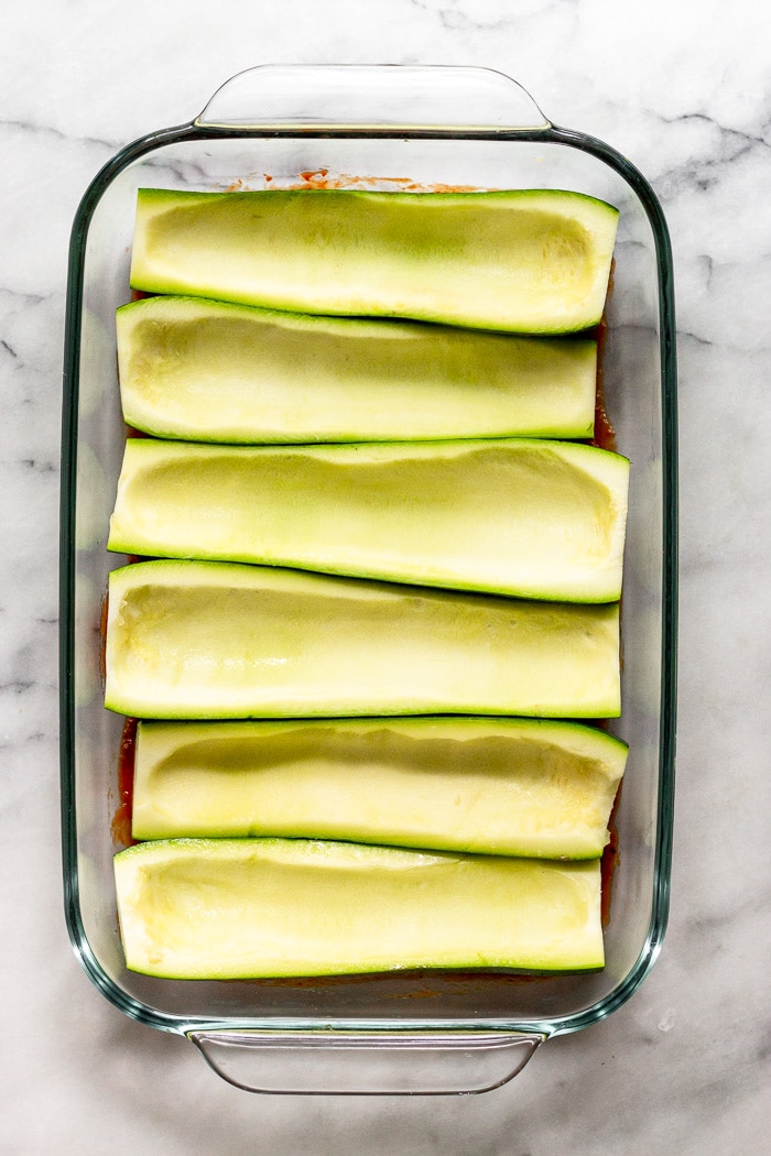 Large glass baking dish filled with raw zucchini boats sitting on top of pizza sauce.