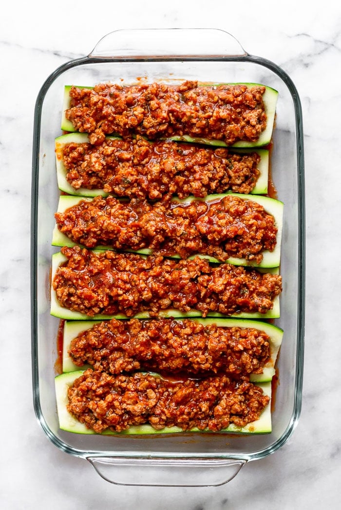 Large glass baking dish filled with raw zucchini boats stuffed with sausage and pizza sauce sitting on top of pizza sauce.