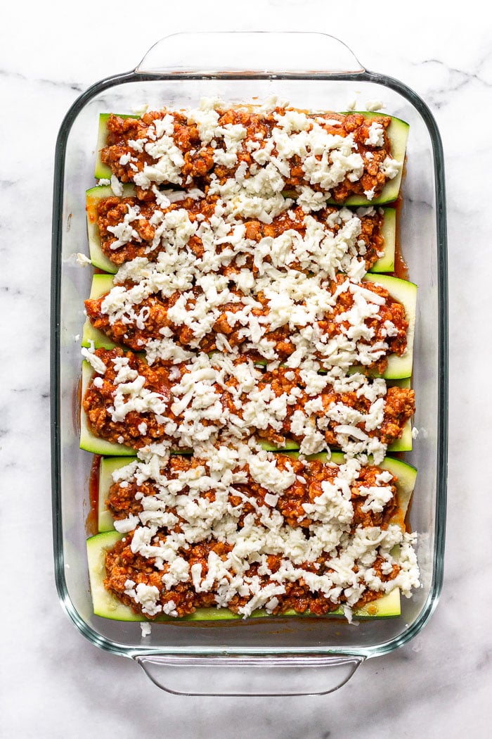 Large glass baking dish filled with raw zucchini boats stuffed with sausage, pizza sauce, and shredded cheese. The zucchini is sitting on top of pizza sauce.