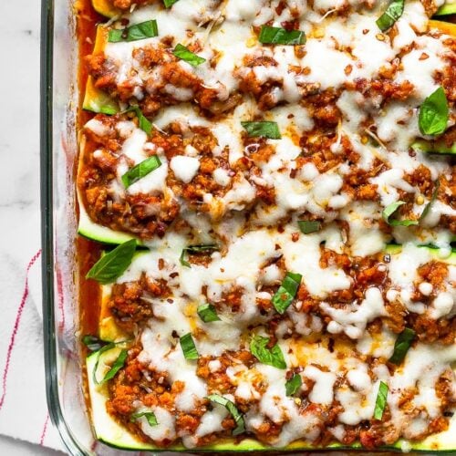 Healthy zucchini pizza boats in a large glass dish toped with fresh basil and red pepper flakes. The pan is sitting on a stripped towel with basil leaves next to it.